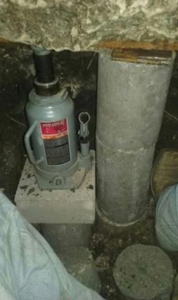 A gray hydraulic bottle jack with red label next to a metal cylindrical container on a concrete floor in a basement or garage