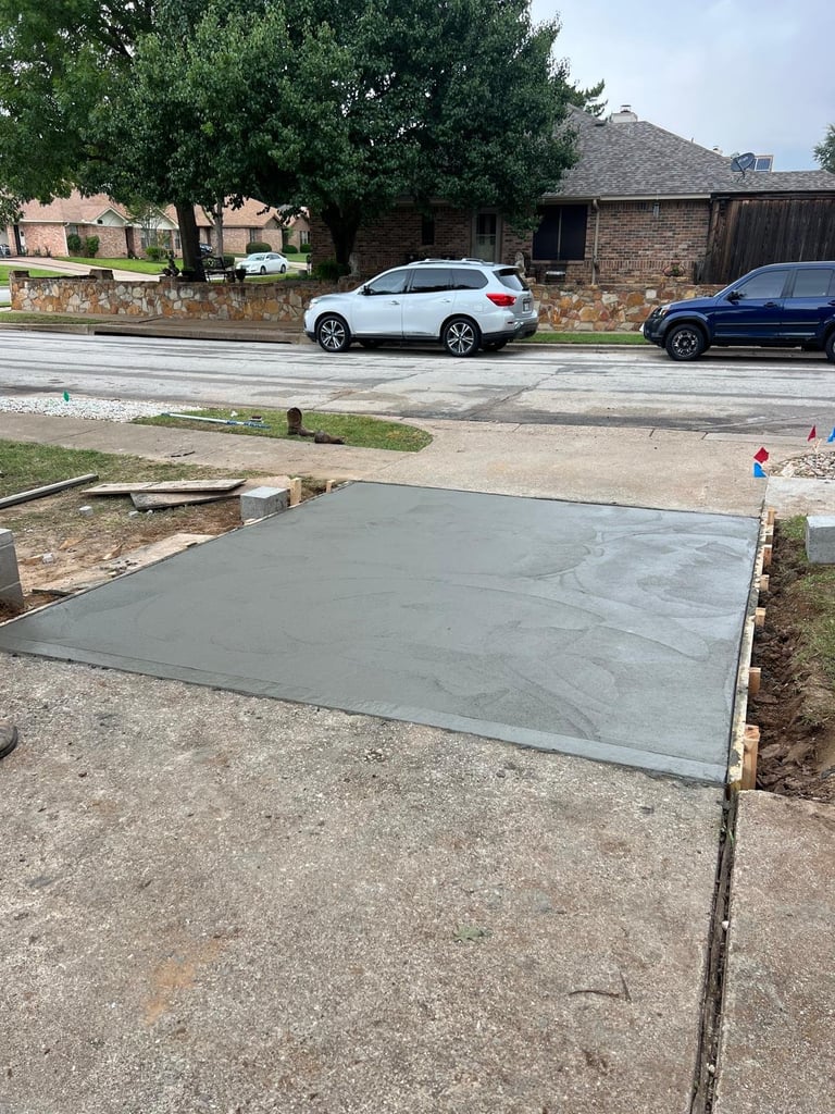 Freshly poured concrete pad in residential driveway with parked vehicles and brick home visible in background